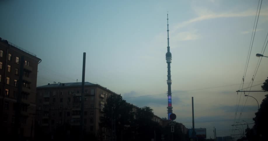 Vehicle follows overhead lines past buildings and railway signals toward tower with spire