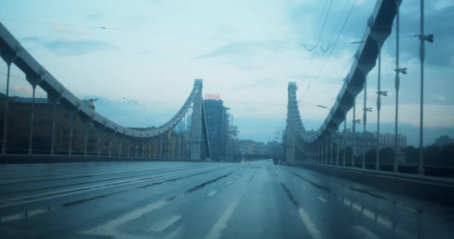 Wet asphalt road leads over bridge to low rise buildings under cloudy sky