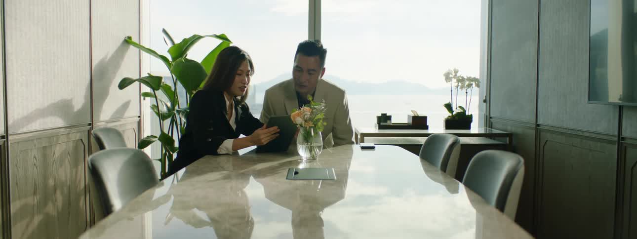 A man and woman sit down in a conference room and look at a laptop