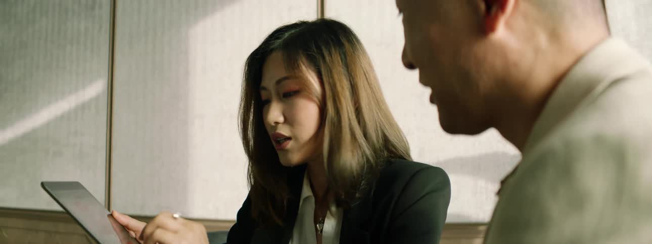 A woman sitting at a table in an office shows a tablet to a man as they speak with one another