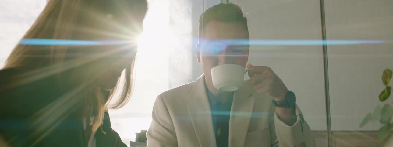 A man and woman sit at an office table with a tablet and the man drinks from a small mug