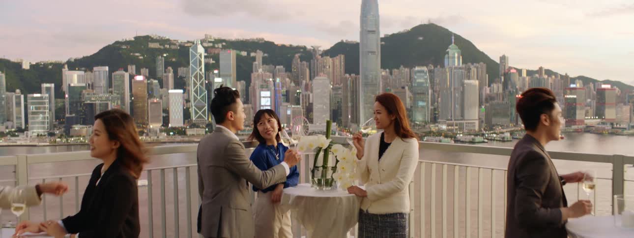 A group of people gather on a rooftop drinking wine beside a water source and large city