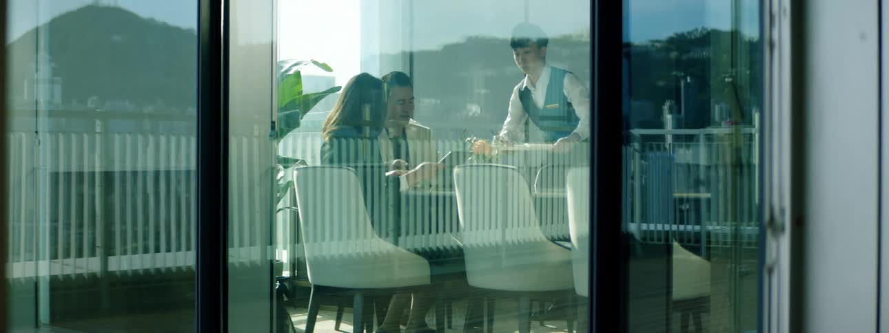 A man and woman sit at a restaurant and look at a menu while a server brings some dishes out