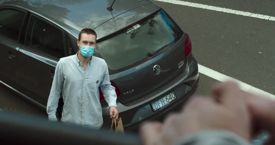 Man wearing face mask gestures with food delivery bag up toward person waiting at balcony rail