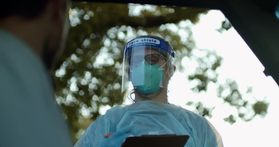 A nurse asks questions to a patient outside a clinic A medical professional wears full protective gear holding a clipboard next to a vehicle at drive through COVID test clinic