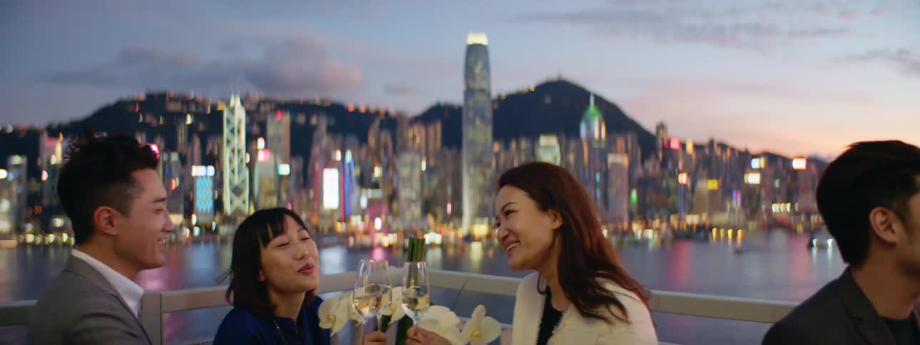 People sitting at a restaurant and knocking champagne glasses with the city skyline the sky and clouds in the background