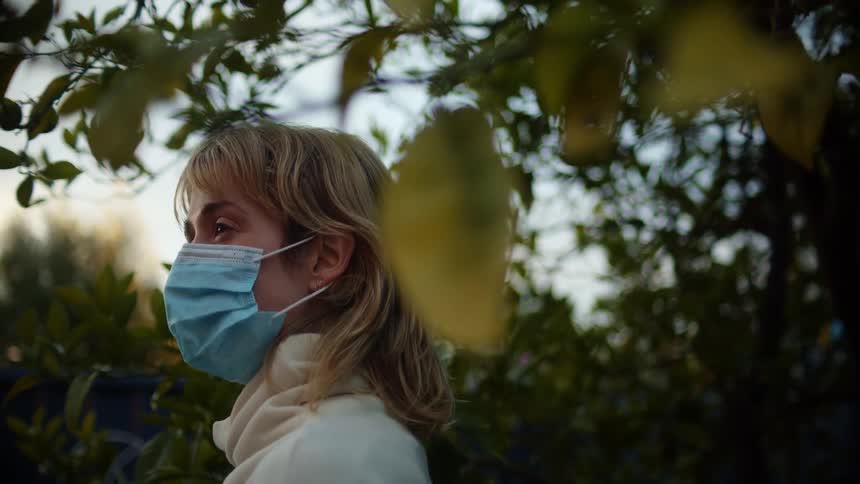 A woman stands next to a large tree outside while wearing a blue disposable face mask