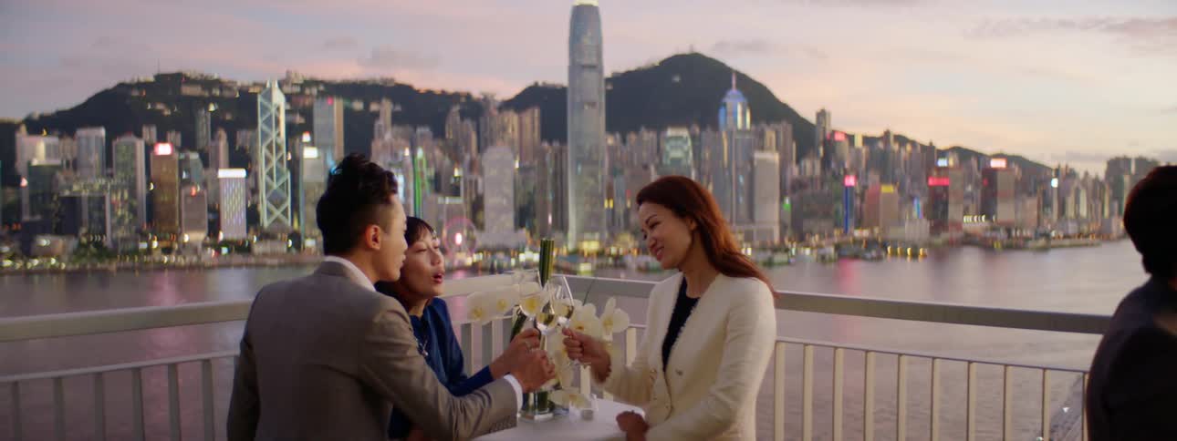 Groups of people toast and drink wine while standing at tables on a balcony overlooking the ocean at sunset