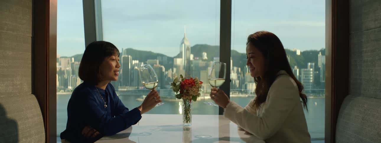 A pair of asian women sitting together in a booth in a restuarant clink their glasses together before taking a sip of their white wine
