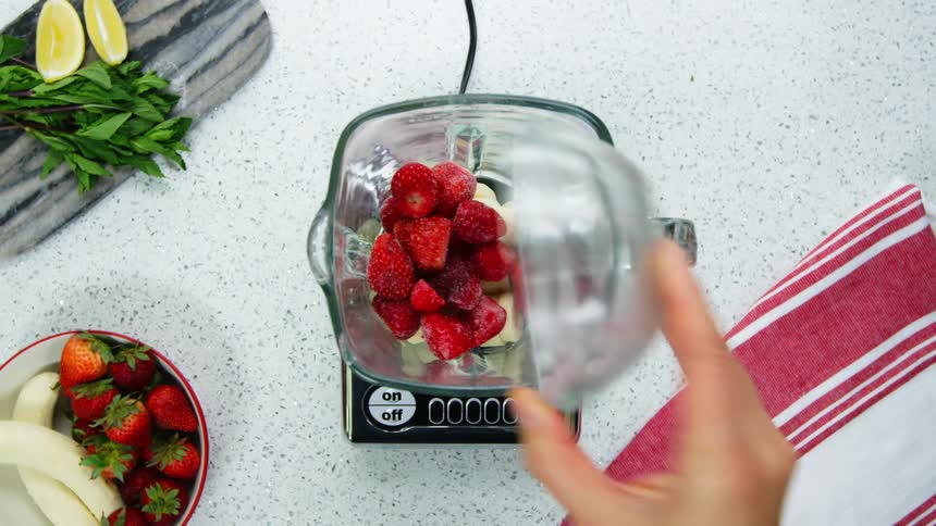 A person pours strawberries and sliced bananas into a blender on a kitchen counter