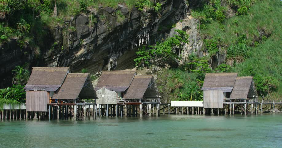 Person walks along dock near hut like homes perched on stilts against rainforest and mountain slope