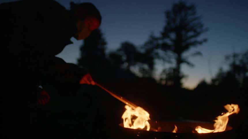 A person wearing a face mask pokes a campfire with a stick at night