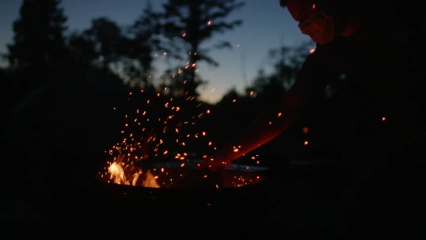 A man wearing a medical mask places a log into a fire in a barrel in a woodland at sunset