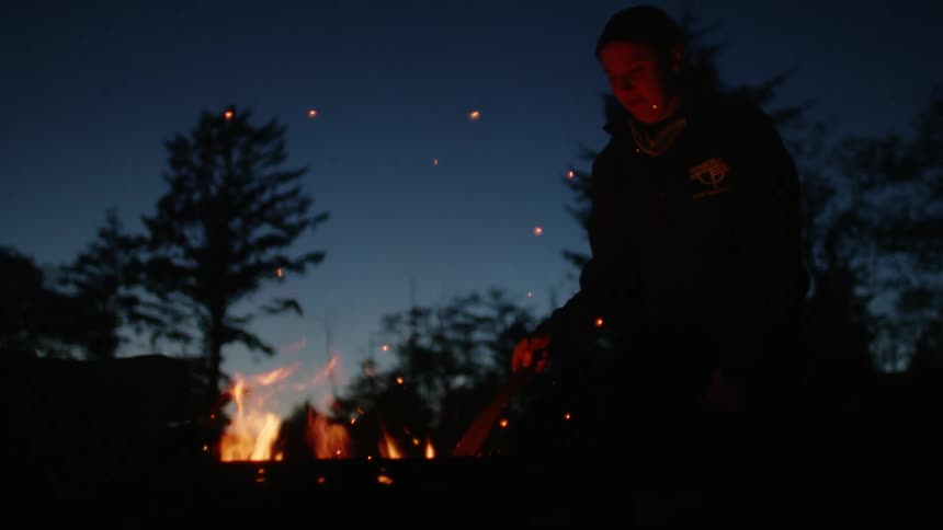A person stands beside a campfire at night and pokes at it with stick
