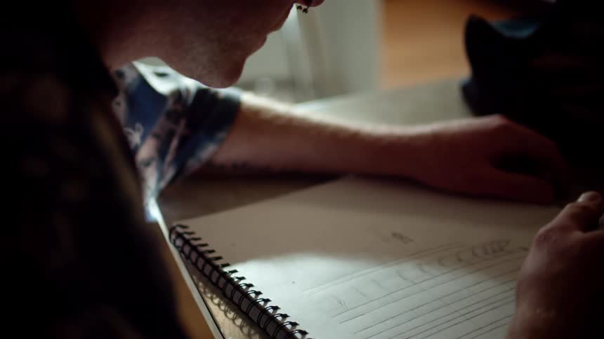 A man sitting at a desk uses a pen to draw in a notebook