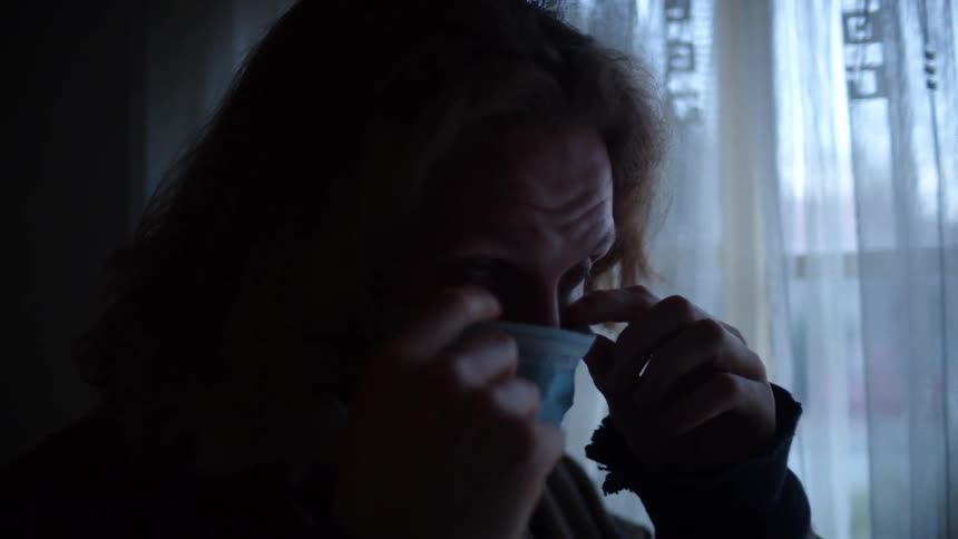 A man standing next to a window in a dimly lit room puts on a medical mask and adjusts his hair