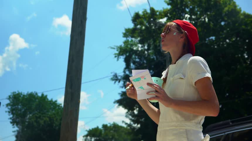 A woman painter wearing a red hat and white overalls and sunshades is standing back from her painting thinking of what to paint next