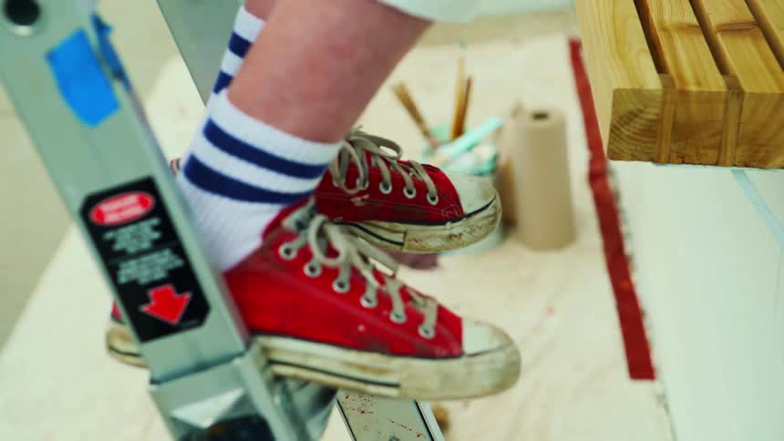 A woman wearing red sneakers standing on an aluminum ladder painting a sign
