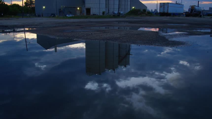 Large puddle in parking lot near factory with tanks reflects clouds as tinted sky sits over skyline on horizon