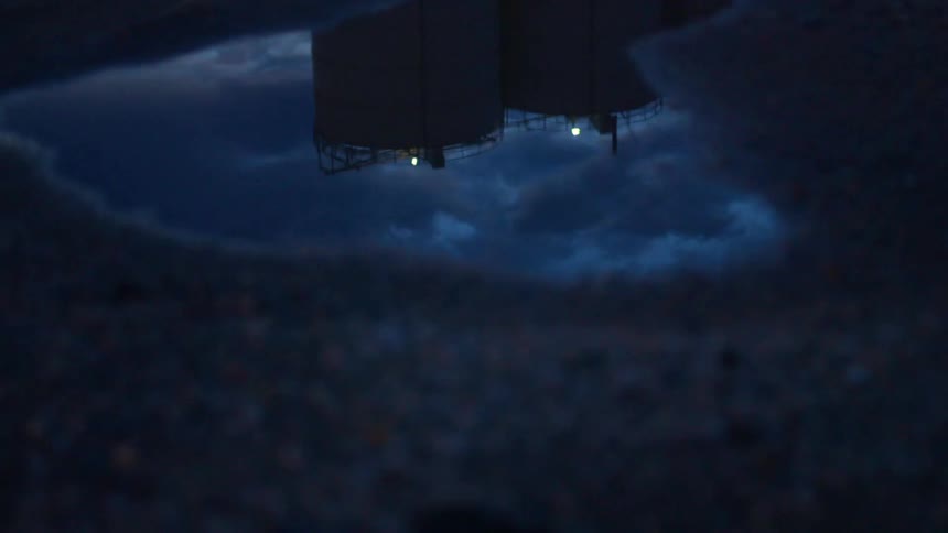 Puddle reflects tanks with rail and lit lights under cloudy sky at night
