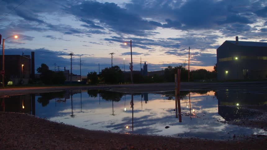 Street lamps and clouds reflected in a puddle near buildings at dusk