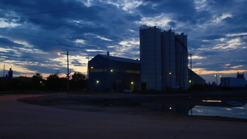 Factory lights glow near building and tanks with semi trailer parked in lot and city skyline visible on horizon