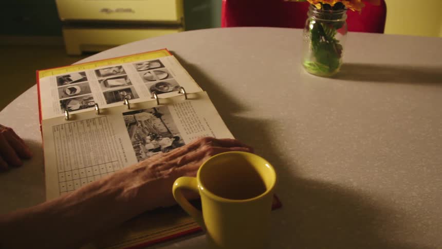 Elderly woman sits at table in kitchen looking through cookbook laying near yellow coffee mug