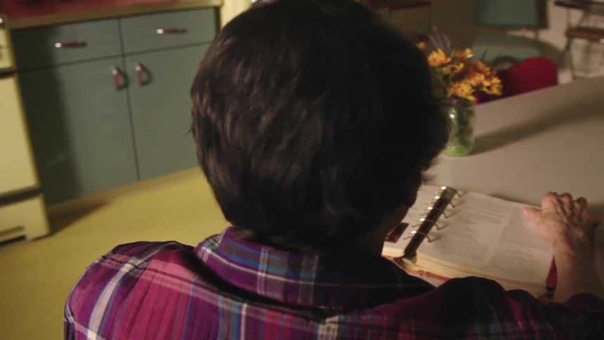 An elderly woman sits at a table in an old fashioned kitchen and flips through a recipe book