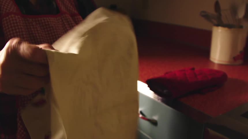 Elderly woman with a plaid shirt and an apron dries her hands with a towel in the kitchen