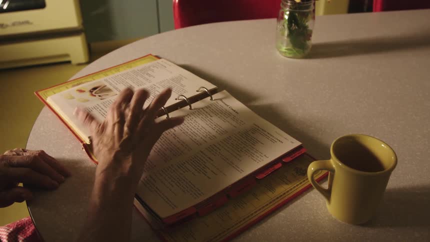 An elderly woman looks through a recipe book while sitting at a table in a kitchen