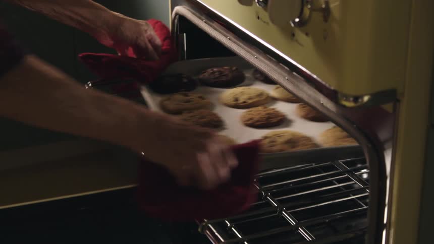 A woman sitting at a table in a kitchen then taking cookies out of a oven