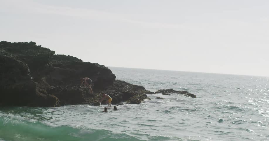 A pair of men climb out of the ocean onto a rock formation while two women watch from the water