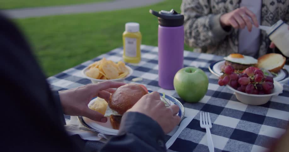 People eat hamburgers topped with fried eggs at picnic table near asphalt path