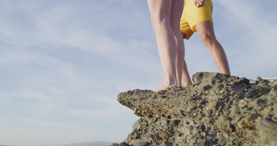 A young man and woman in swimsuits approach the edge of a rock formation over the ocean and prepare to dive