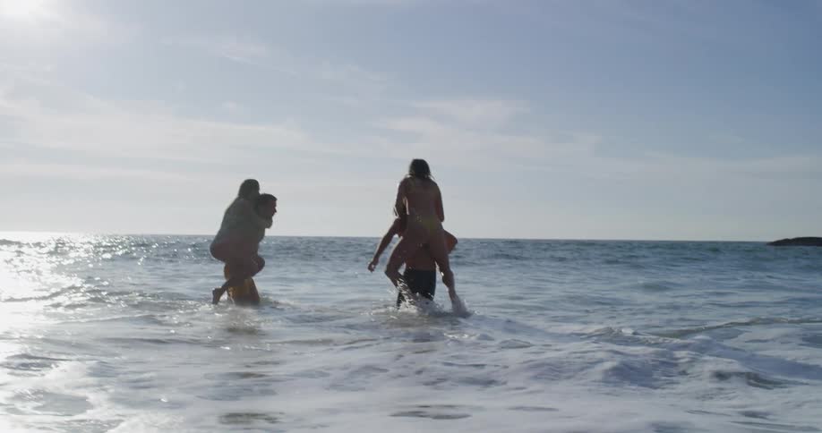 A pair of women climb onto the backs of a pair of men standing in the tide on the beach during the day