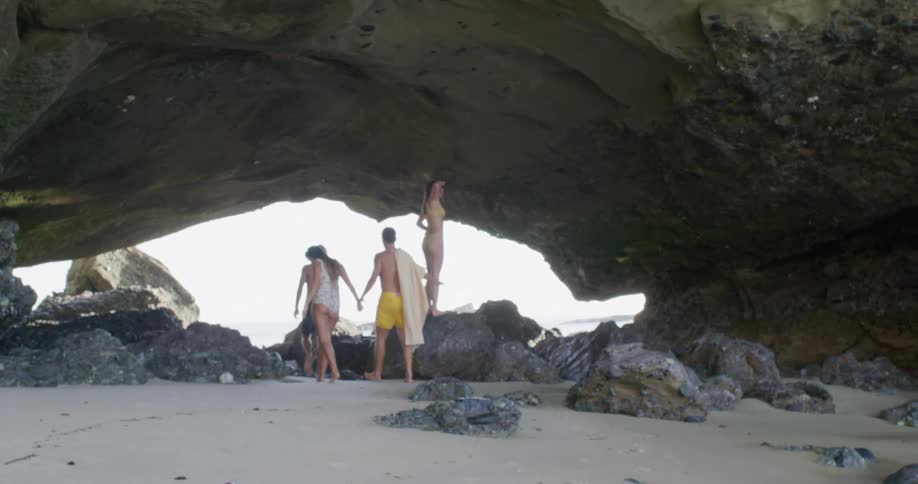 A group of men and women in swimsuits walk through a cave on a beach during the day