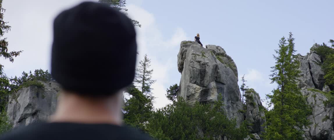 A man standing on the ground stares at a woman sitting on the top of a tall rock in a canyon
