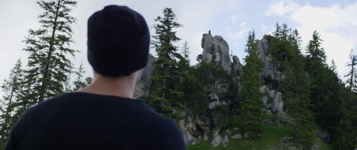 A man walking outdoors on a sunny day stops to look up at a mountain and the evergreen trees that surround it