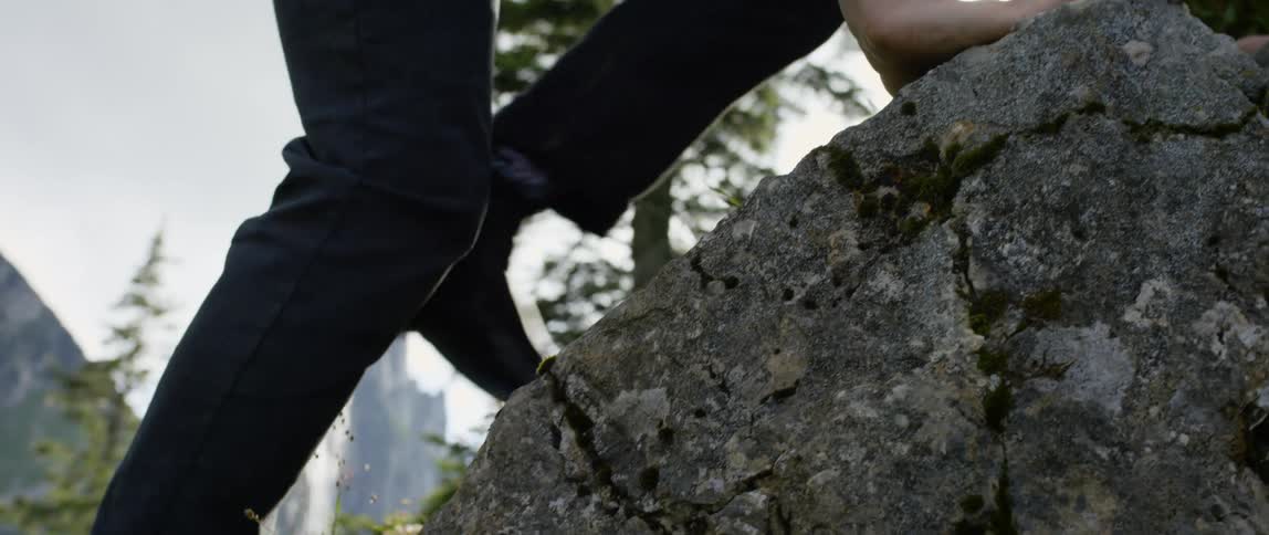 A man dressed all in black uses his hands and feet to climb over boulders on the side of a hill
