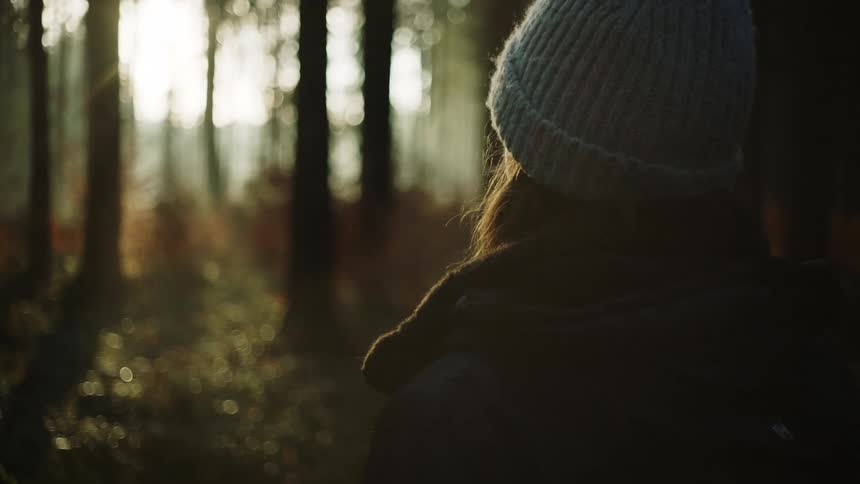 A woman wears a thick coat and a knit hat as she stands in a forest