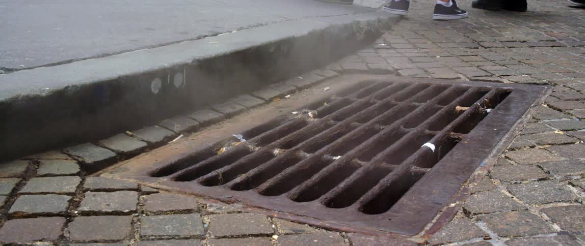 Steam rises from a sewer grate next to a sidewalk on a cobblestone street