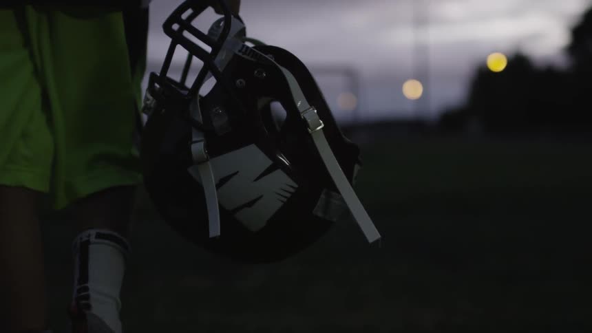 A boy walking across a field while carrying a football helmet