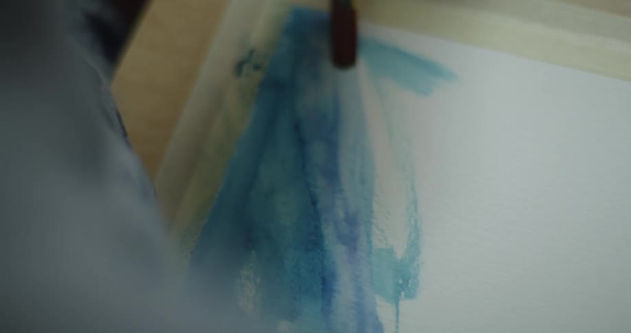 A person paints with blue watercolor over paper with a small brush