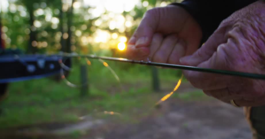 Older man strings line onto fishing pole near truck parked on dirt road as sun rises