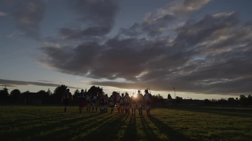 A group of people carrying weapons walk along a field at dawn