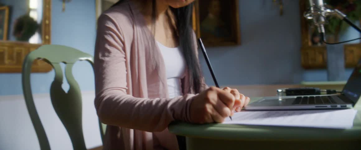 A woman sits at a table with a laptop and writes on a piece of paper