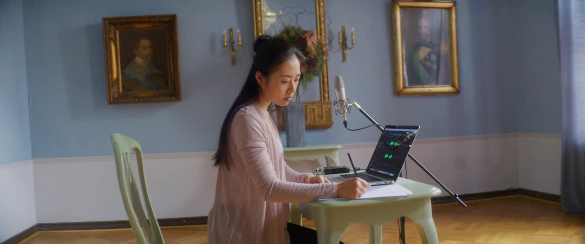 Woman sits at table near microphone and laptop showing audio related display writing on paper in room with framed art