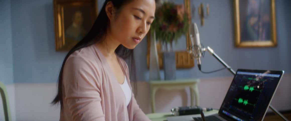 Woman wearing cardigan sits near laptop and microphone writing on musical sheet