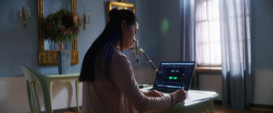 Woman sits at table writing in ornately decorated room near microphone and laptop showing sound software gui