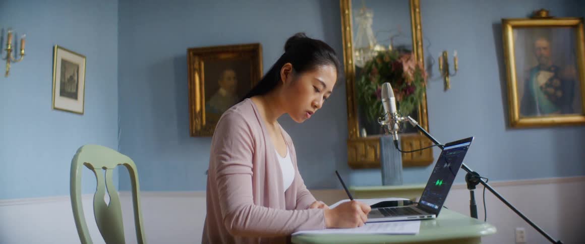 Woman writes on paper sitting at table near laptop and microphone next to wall with gold framed art